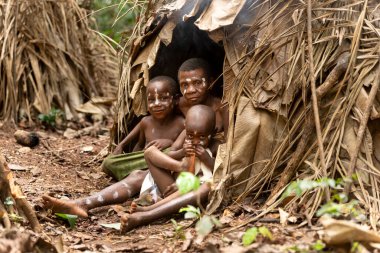 Bir pigme adam, Kamerun ormanının ortasında, geleneksel bir yaprak kulübesinin önünde iki çocukla oturuyor. Bir çocuk palayla oynarken diğeri büyükler ona sarılıyor..