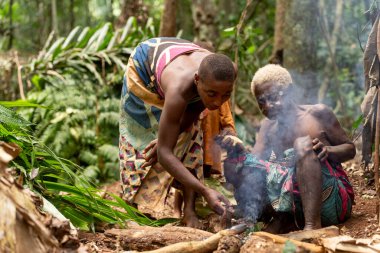 Baka kabilesinden bir pigme, Kamerun ormanındaki ateşin yanında yaşlı annesine yardım ediyor. Bir anlık bakım ve geleneksel günlük yaşam