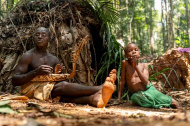Yaşlı bir Baka adamı, yağmur ormanlarında çocuklarla ve kabile üyeleriyle çevrili geleneksel bir telli çalgı çalar.