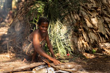 Gülümseyen Pigme kadın tropikal ormanda kulübenin önünde ateş yakıyor, geleneksel orman yaşamının otantik anı.