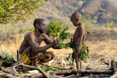 Dupa kabilesinden Kamerunlu bir adam, genç bir çocuğa geleneksel bir yaprak etek hazırlamasında yardım ediyor, ona kırsal Afrika doğasında kültürel uygulamaları öğretiyor.