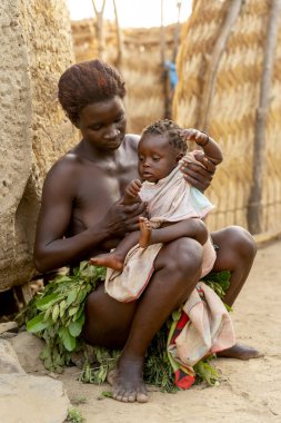 Kamerun 'daki Dupa kabilesinin annesi bebeğini kucağında, köy ortamında geleneksel bir yaprak etek giyiyor. Otantik Afrika günlük yaşamı