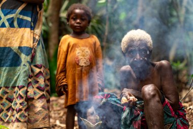 Yaşlı bir Baka kadını ateşin yanında bir çocukla oturuyor. Nesillerin ve geleneksel orman hayatının güçlü bir portresi..