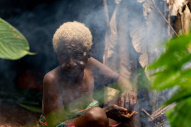 Yaşlı Baka kadın kulübesinde duman içinde oturuyor. Buruşuk yüzü orman yaşamını ve kabile mirasını yansıtıyor..