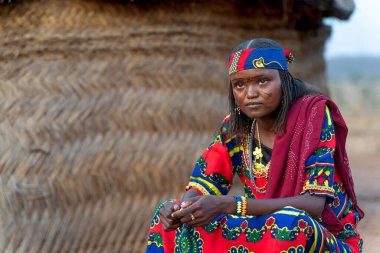 Yüzünde dövme olan renkli geleneksel elbiseli Mbororo kadın otantik güzelliği ve kırsal Kamerun geleneklerini yansıtan bir kulübede oturuyor..