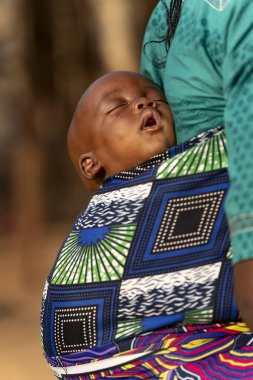 Uyuyan Mbororo bebeği, anne sırtına renkli bir kumaş sarılmış. Afrika kültüründe barışı, güvenliği ve anneliğin derin bağını simgeliyor..