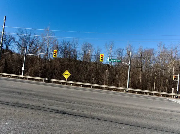 11. Otoyol 'da kırmızı ışık ve yol işaretleri olan bir kavşak. Sarı bir tabela sürücüleri potansiyel bir bölünme veya dönüş konusunda uyarırken, ana yol tabelası otoyolu tanımlıyor. Çevredeki çevre sakin bir kırsal alan olduğunu gösteriyor.