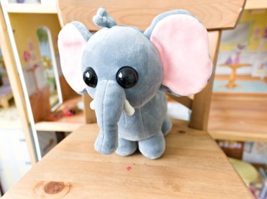 Gri filli peluş bir oyuncak ahşap bir sandalyede oturuyor. Sandalye bir sürü mobilyayla dolu bir odada, yemek masası ve birkaç kitap da dahil.