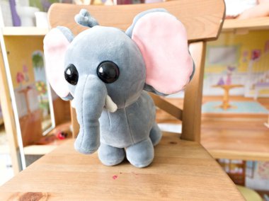 Gri filli peluş bir oyuncak ahşap bir sandalyede oturuyor. Sandalye ahşap zemini ve ahşap kitaplığı olan bir odada.