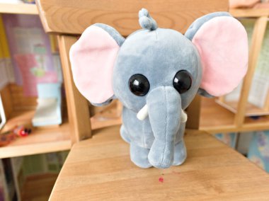 Gri bir fil oyuncağı ahşap bir sandalyede oturuyor. Filin pembe bir sandığı var ve kameraya bakıyor.