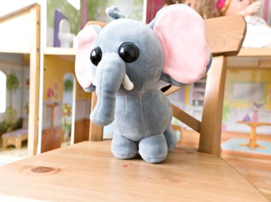 Doldurulmuş bir fil ahşap bir sandalyede oturuyor. Filin pembe bir kulağı ve siyah bir burnu var. Sandalye bir bebek evinin önüne konur.