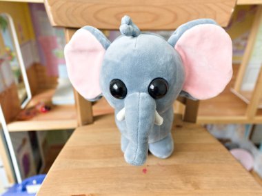 Gri bir fil oyuncağı ahşap bir sandalyede oturuyor. Filin pembe bir hortumu ve pembe bir kulağı var.