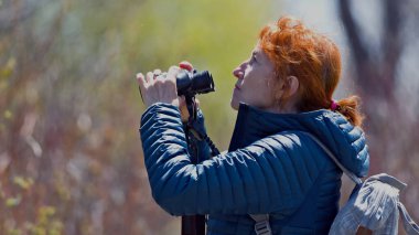 Mavi ceketli ve kırmızı atkuyruklu bir kadın dürbünden bakıyor.