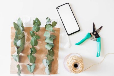 Cep telefonu, el işi kağıdı ve beyaz arka planda okaliptüs dalları olan minimal çiçekçi çalışma alanı  