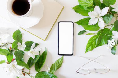 İçi boş cep telefonu ve fotokopi alanı olan ofis masası. Beyaz çiçekler ve böğürtlen yaprağı çerçevesi. Bardaklar, bir fincan kahve ve beyaz arka plan üzerine açık bir kitap. Düz yatıyordu. Bahar Kompozisyonu. 