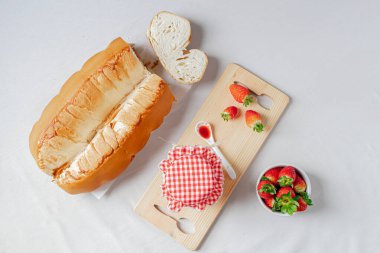 Beyaz keten kumaş arka planda bir kavanoz çilek reçeli, bir kase taze çilek ve lezzetli bir ekmek..  