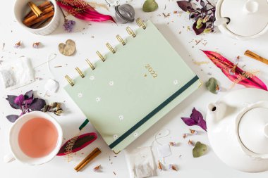 Çay saati kadınsı çerçeve kompozisyonu, bir planlayıcı, çay demliği, tarçın, fincan, çay süzgeci, mor ve pembe yapraklar ve beyaz arka planda kurumuş bitkiler. Düz yatış, üst görünüm.