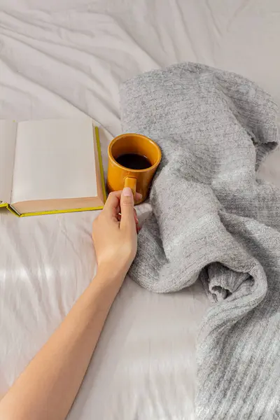 一个女人在床上拿着一杯咖啡 拿着一本书 穿着舒适的毛衣 自由自在地在家里干活 懒洋洋的 舒适舒适的家的概念 — 图库照片