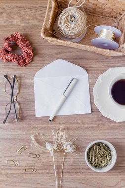 Tahta masadaki zarf. Ofis masası çalışma alanı, yabani çiçekler, bir fincan kahve, saç tokası ve kırtasiye malzemesi. Düz yerde, üst tarafta. Samimi kompozisyon. Romantik harf kavramı. 