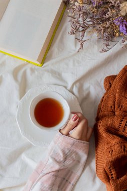 Bir fincan çay tutan genç bir kadın, bir süveter ve bir kitaba yakın. Autumn, sonbahar kompozisyonu. Samimi konsept. 