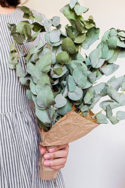 Beyaz arka planda okaliptüs buketi olan kamış sepeti tutan çizgili, klasik elbiseli genç ve güzel bir kadın. Çiçek asgari konsept.