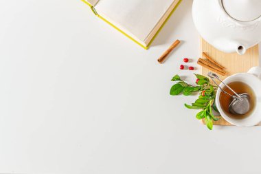Çay süzgeci, çaydanlık, açık kitap, tarçın, beyaz arka planda yeşil yapraklar. Düz yatış, üst görünüm.