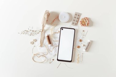 Dikiş araçlarıyla boş kopyalama alanı: düğmeler, fermuar, mezura, iplikler, iğneler ve beyaz arkaplanda akıllı telefon. Düz yerde, üst tarafta. Hobi, eğlence konsepti. 