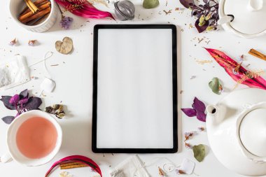 Çay saati çerçeve bileşimi dijital tablet ve çaydanlık, tarçın, fincan, çay süzgeci, mor ve pembe yapraklar ve beyaz zemin üzerinde kurumuş bitkiler. 