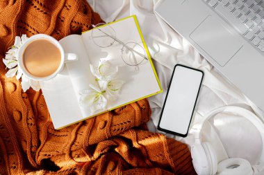 Yavaş sabah kompozisyonu. Kazak, kahve ve süt, çiçekler, akıllı telefon, kitap ve kolye dağınık bir yatakta. Sonbahar, sonbahar konsepti.