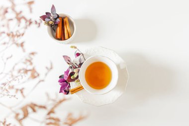 Bir fincan çay, tarçın ve mor yapraklar beyaz arka planda. Düz yatış, üst görünüm.