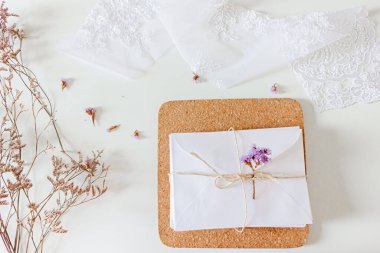 Bej rengi düğün davetiyeleri kompozisyonu. Beyaz zarf, kurumuş çiçekler, dantel kumaş, beyaz arka planda sisal ip. Düz yerde, üst tarafta. Nötr durağan. 