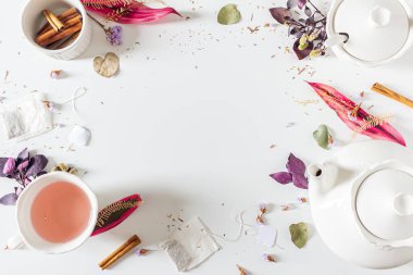 Çaydanlık, tarçın, fincan, çay süzgeci, mor ve pembe yapraklar ve beyaz arka planda kurumuş bitkilerle çay zamanı çerçevesi bileşimi. Düz yatış, üst görünüm. 