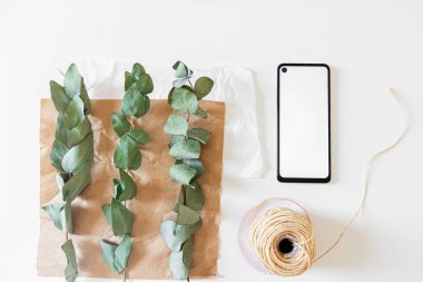 Cep telefonu, el işi kağıdı ve beyaz arka planda okaliptüs dalları olan minimal çiçekçi çalışma alanı  