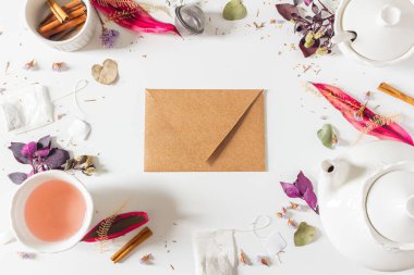 Çay saati kadınsı çerçeve kompozisyonu Kraft zarfı, çay demliği, tarçın, fincan, çay süzgeci, mor ve pembe yapraklar ve beyaz arka planda kurumuş bitkiler. Düz yatış, üst görünüm. 