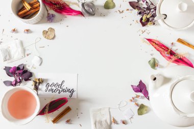 Çaydanlık, tarçın, fincan, çay süzgeci, mor ve pembe yapraklar ve beyaz arka planda kurumuş bitkilerle çay zamanı çerçevesi bileşimi. Düz yatış, üst görünüm. 