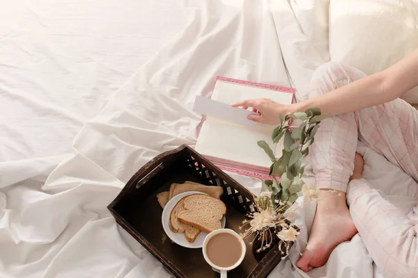 女人躺在床上 用深色的木制复古托盘阅读一本书和早餐 上面有米色的床单和枕头 — 图库照片