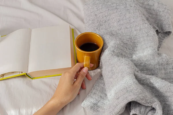 一个女人在床上拿着一杯咖啡 拿着一本书 穿着舒适的毛衣 自由自在地在家里干活 懒洋洋的 舒适舒适的家的概念 — 图库照片