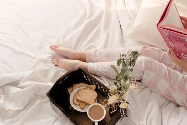 女人躺在床上 用深色的木制复古托盘阅读一本书和早餐 上面有米色的床单和枕头 — 图库照片