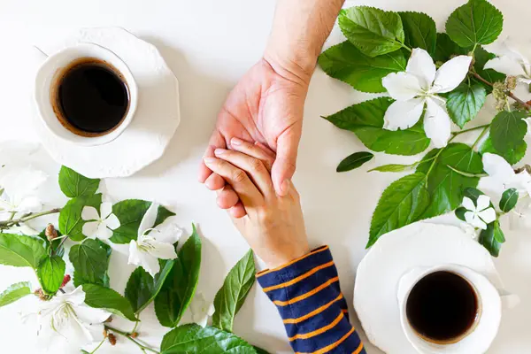 Woman and man holding hands, white flowers and green leaves frame and two cups of coffee. Flat lay, top view. Spring composition. Romance concept.