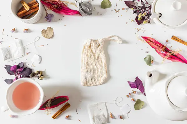 Tea time frame composition with cotton, bag, tea pot, cinnamon, cup, tea strainer, purple and pink leaves and dried plants on white background. Flat lay, top view.