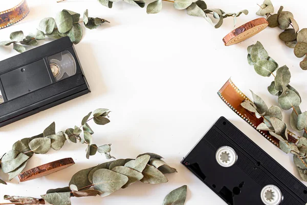 Video cassette, old-fashioned camera films, dried branches of eucalyptus leaves, and wood pieces on white background. Flat lay, overhead view, top view. Vintage frame composition.
