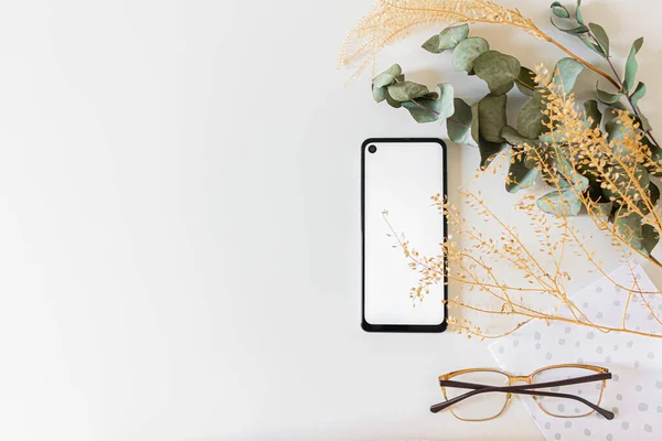 mockup image of black and white mobile phone and black blank paper sheet with eucalyptus branches on white background. flat lay.