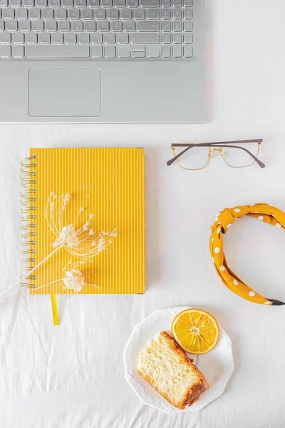 Home office desk with laptop, orange piece of cake, headband, glasses, beige wildflowers and notebook on white background. Flat lay, top view. Feminine business concept.