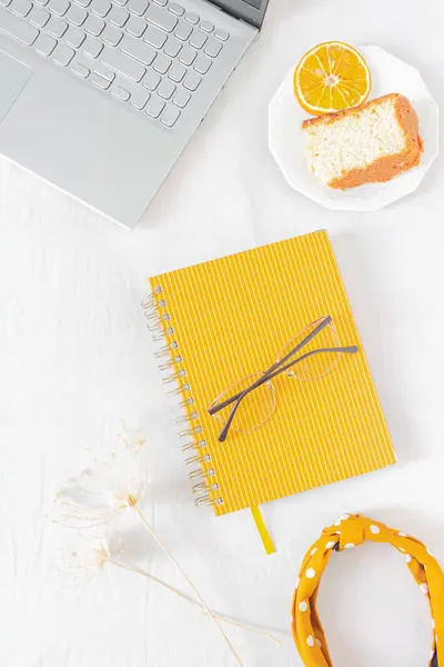 Home office desk frame with laptop, orange piece of cake, headband, glasses, beige wildflowers and notebook on white background. Flat lay, top view. Feminine business concept.