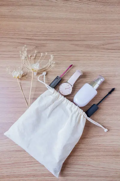 Female styled accessories: cotton bag, wrist watch, foundation, mascara and lipstick on wooden background. Flat lay, top view trendy feminine background.