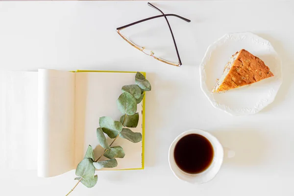 Breakfast composition with cup of coffee, cake slice, eyeglasses and book on white background.