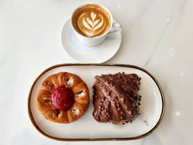 Beyaz bir tabakta ve beyaz arka planda Blossom Croissant ve Chocolate Rocher Croissant 'lı bir fincan sıcak latte sanat kahvesi.