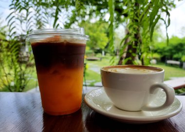 Buzlu siyah portakallı kahve ve ahşap bir masada bir fincan sıcak latte sanat kahvesi ve bulanık bahçe arka planı.