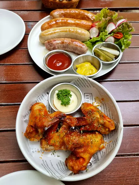 Barbecue sauce baked chicken wings with tartar sauce and a set of German sausages and salad with tomato and white cream sauce on a wooden table.