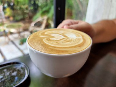 Sağ elin bir fincan sıcak latte sanat kahvesini bulanık, rahat bir bahçe arka planında tutuşu.